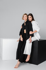 brunette woman hugging friend wearing black clothes while sitting on cube on grey background.
