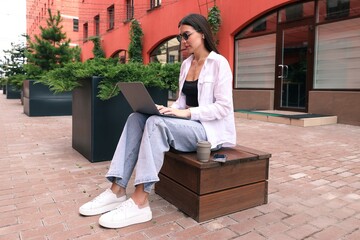 A young woman is sitting on a bench and working with a laptop on the street © makedonski2015
