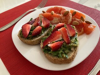 On the table is a white plate on which are laid two crispy toast with a sauce of greens. The toast is garnished with slices of strawberries. Slices of grapefruit with almond nuts lie next to it.