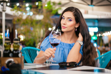 A young woman with bright makeup drinks wine from glass in a street cafe, looking into the camera