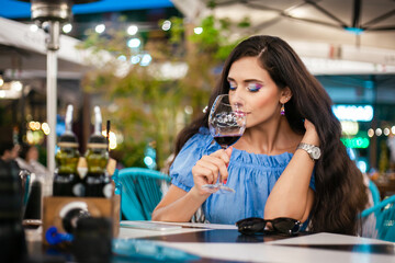 A young woman with bright makeup inhales the aroma of wine from a glass in a street cafe