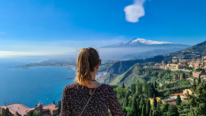 Tourist woman with panoramic view on snow capped Mount Etna volcano and the Mediterranean coastline on a sunny day seen from ancient Greek theater of Taormina, island Sicily, Italy, Europe, EU. Awe - obrazy, fototapety, plakaty