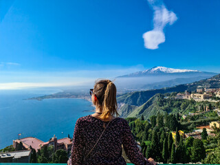 Fototapeta na wymiar Tourist woman with panoramic view on snow capped Mount Etna volcano and the Mediterranean coastline on a sunny day seen from ancient Greek theater of Taormina, island Sicily, Italy, Europe, EU. Awe