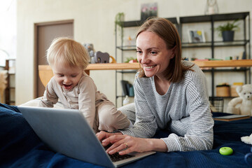 Smiling young mother typing on laptop and doing her online work while her child playing next to her on bed