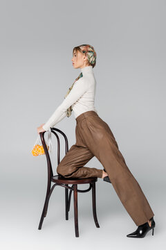 side view of woman in trousers posing with chair and mesh bag with oranges on grey background.