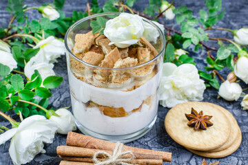 Romantic breakfast. Multi-layer cottage cheese dessert with yogurt and cookies, sprinkled with cocoa in a glass glass, cinnamon sticks, anise, cracker, bushy white roses on a black (dark) background.