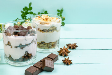 Multi-layer cottage cheese dessert with yogurt, chocolate corn balls and chocolate slices, banana dessert with cookies in a glass glass on a blue (mint) background. Space for the text. 