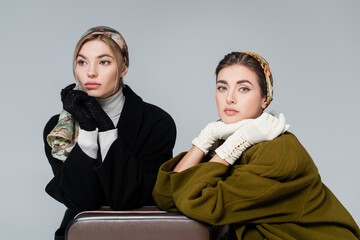 elegant woman in gloves looking at camera near friend and retro suitcase isolated on grey.