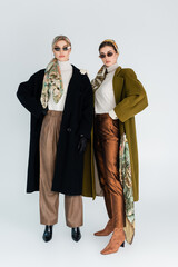 full length of women in trendy coats and sunglasses standing on grey background.