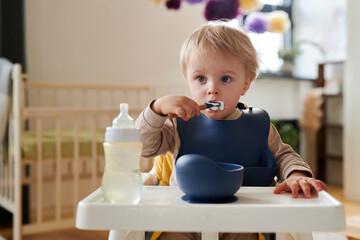 Cute blonde little boy in bib eating porridge with spoon sitting in his chair - obrazy, fototapety, plakaty