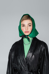 stylish woman in black leather jacket and green kerchief looking at camera isolated on grey.