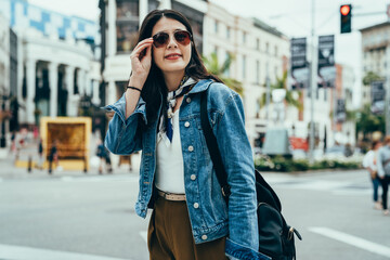 Fototapeta premium asian woman in smart casual is touching her sunglasses and smiling with confidence. blurred background happy female tourist standing at the roadside and looking into distance