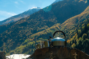 Close-up of a metal teapot and two cups on a stone against the background of mountains on a sunny day, copy the space. Beautiful, atmospheric autumn landscape of picnic mountains