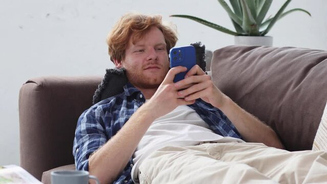 Young redhead man using mobile phone, chatting or playing games while lying on the couch at home. Relaxing at home, smiles friendly