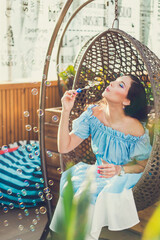 A young woman with bright makeup is sitting in a summer outdoor cafe in a hanging chair and blowing soap bubbles