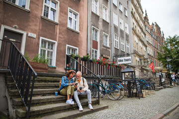 Fototapeta na wymiar Happy senior couple tourists sitting on stairs and having take away coffee outdoors in town