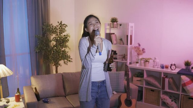 taiwanese female having a spree singing karaoke and drinking alcohol in apartment. asian woman with microphone lifting beer bottle up in the air to celebrate the upcoming long national holiday.