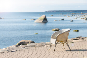 Fototapeta na wymiar Beach cafe chair with blurred sea background. Summer vacation, travel, holidays concept. Lonely, empty, white wicker seat on shore pavement in sunshine. Seaside restaurant on sunny day. Soft focus
