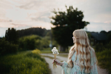 young long-haired blonde in a dress with dandelions in her hands stands at sunset in the village.