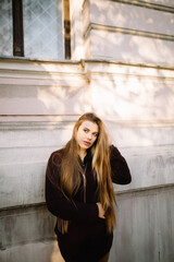 Fototapeta premium Woman with long hair and big lips stands near the wall of an old city building, touches her hair and looks away. Light effect in the sun.