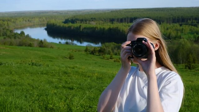 A fair-haired girl photographs nature on a summer day. The girl is facing the operator. The girl takes pictures with a digital camera.