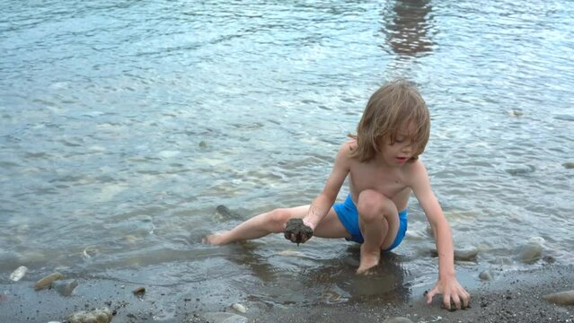 Cute little boy with long blond hair playing with sand nearby the see.
