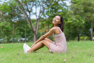 Naklejka na ściany i meble Portrait photo of a young beautiful asian female lady in pink dress sitting on the grass embracing the breeze wind and soft sunlight relaxingly and chillingly in a public park
