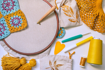 Flatlay of embroidery hoop with blank fabric and punch needle inserted in, crochet hook, tassels, heart, spool of thread, marker and crochet patterns on white surface. © Uliana