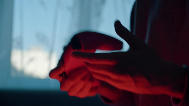 Close-up of young male hands holding gamepad, playing video game, pressing buttons, controller stops working due low battery, man shakes and knocks on broken device. Dark red room, window daylight.