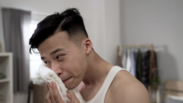 attractive asian man in undershirt walking near the mirror to look at his reflection and wiping face with a towel while getting ready for a new day at home.