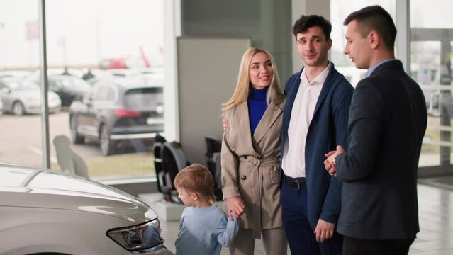 dealership, happy young male and female buyers with a child choose a family car in a showroom and consult with a manager about a new auto model