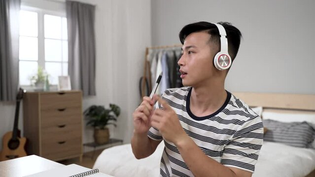 distracted asian young man learning from home is playing with pen like drum stick while listening to pop songs from headphones at desk in the bedroom.
