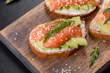 Toast sandwich with butter, avocado and salmon, decorated with arugula and sesame seeds, on a black stone background