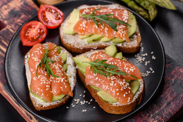 Toast sandwich with butter, avocado and salmon, decorated with arugula and sesame seeds, on a black stone background