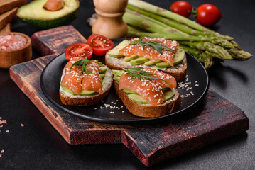 Toast sandwich with butter, avocado and salmon, decorated with arugula and sesame seeds, on a black stone background