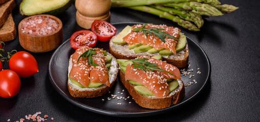 Toast sandwich with butter, avocado and salmon, decorated with arugula and sesame seeds, on a black stone background