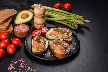 Fototapeta na wymiar Toast sandwich with butter, avocado and salmon, decorated with arugula and sesame seeds, on a black stone background