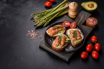 Fototapeta na wymiar Toast sandwich with butter, avocado and salmon, decorated with arugula and sesame seeds, on a black stone background