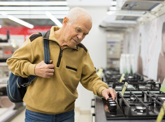 elderly grayhaired man pensioner looking gas hob at counter in showroom of electrical appliance...