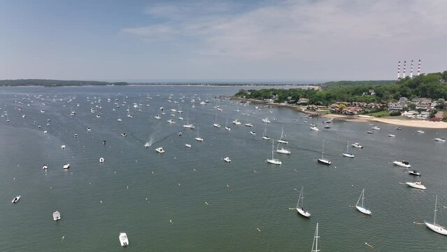 An aerial view over the Northport Marina on Long island, NY with many anchored boats. It is a sunny day with blue skies and white clouds above. The camera dolly in over the sailboats.
