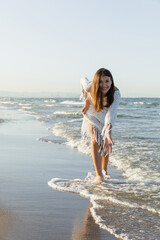 Happy woman in dress looking at camera while standing in sea water on sandy beach.