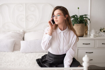A happy emotional European girl in a white shirt and glasses is talking on the phone. Sitting on a white bed