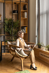 An elderly business woman in a beige trouser suit works in an office sitting in a chair. Working at a computer, typing on a laptop.Senior