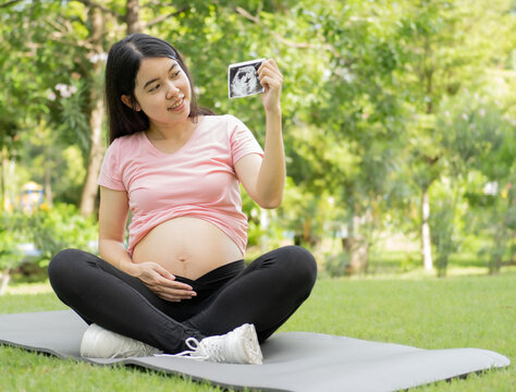 Happy pregnant woman sits relaxing outside in the park looking at an ultrasound image and smiling. Young Asian pregnant woman touching her belly with care and love. Pregnancy and motherhood concept.