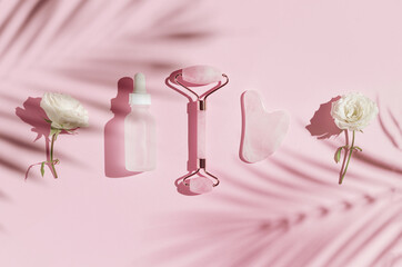 Creative composition made of face roller, gua sha massager, bottle of cosmetic serum on pink pastel sunlit background with white rose buds and shadows. Natural treatment concept. Top view. Flat lay