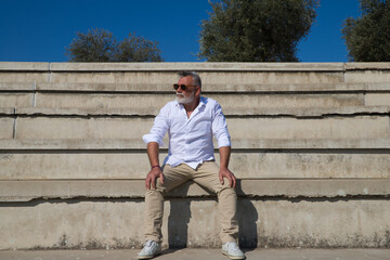 Fototapeta premium handsome mature man with beard and grey hair is sitting on the steps of an open-air auditorium. The man looks at different places. Senior concept, travel and tourism.