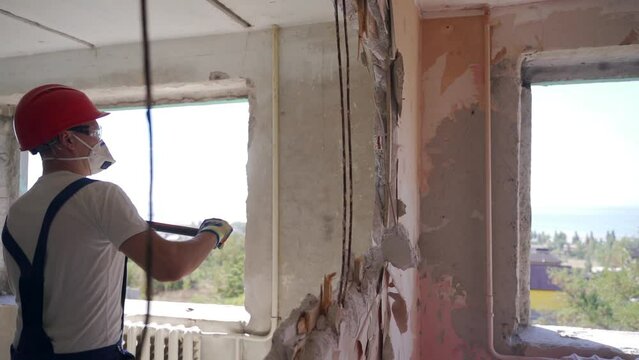 Contractor wrecks wall with sledgehammer making hole for rearrangement. Construction worker doing manual dismantling and demolition works with big hammer hits for apartment renovation in slow motion.