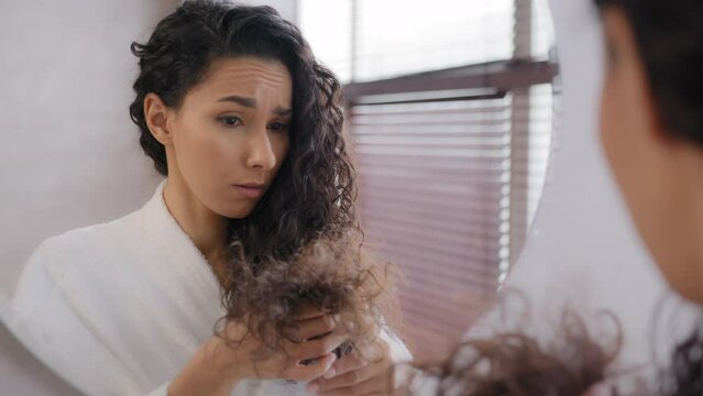 Upset sad young woman distressed by damaged hair frustrated about split ends girl worried about loss brittle dry hair hormone problem vitamin deficiency reflecting in mirror haircare treatment concept