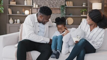 Biracial family sitting on couch in living room spending weekend together. African mother and father with child girl having fun at home. Dad tickling kid daughter on sofa laughing tickle play game