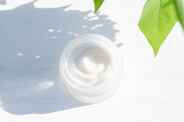 Obraz na płótnie Canvas close up of collagen cream in glass jar and green leaves on white wooden background. Hursh shadows. skin and body care beauty products